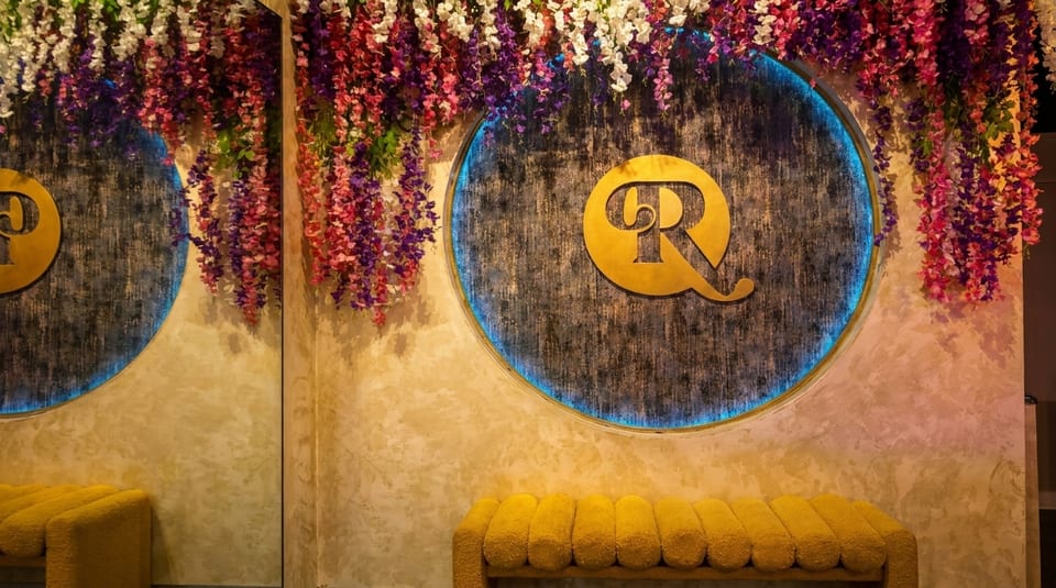A golden "RQ" logo on a dark circular sign with blue backlighting, surrounded by colorful hanging flowers.