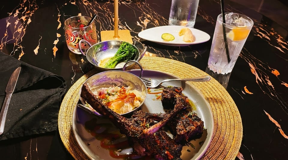 Grilled lamb chops with mashed potatoes, broccolini, and drinks on a dark marble table.