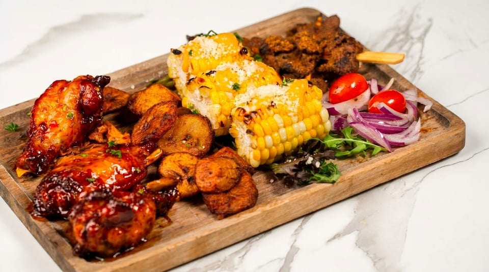 A wooden platter with glazed chicken wings, fried plantains, grilled corn, and a side salad.