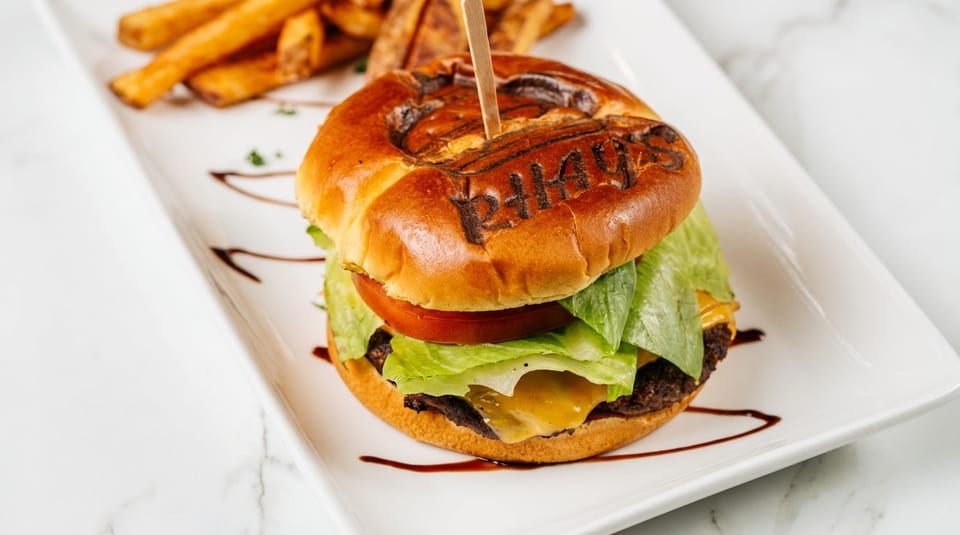 A cheeseburger with lettuce, tomato, and a branded bun, served with fries on a white plate.