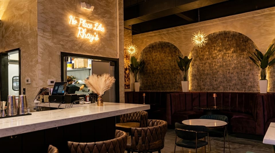 Modern restaurant interior with a bar, neon sign, plush seating, and decorative plants.