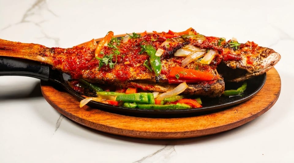 Whole grilled fish topped with red sauce, green and red peppers, and onions on a sizzling platter.